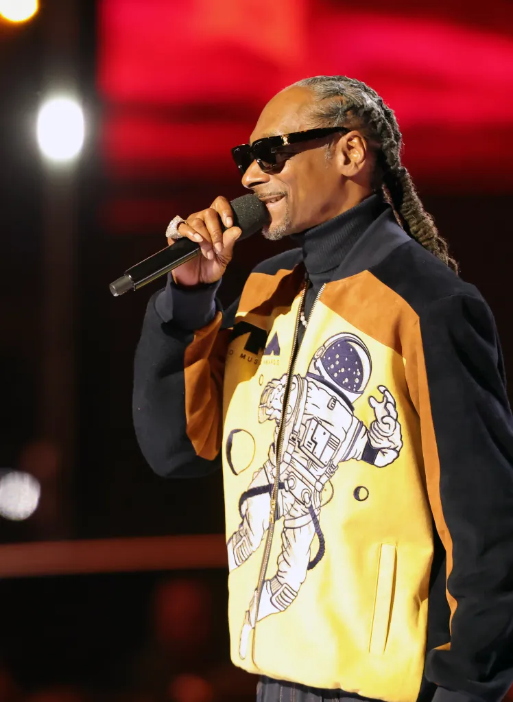 Snoop Dogg speaking into a microphone while wearing sunglasses and a yellow and black jacket with an astronaut design.