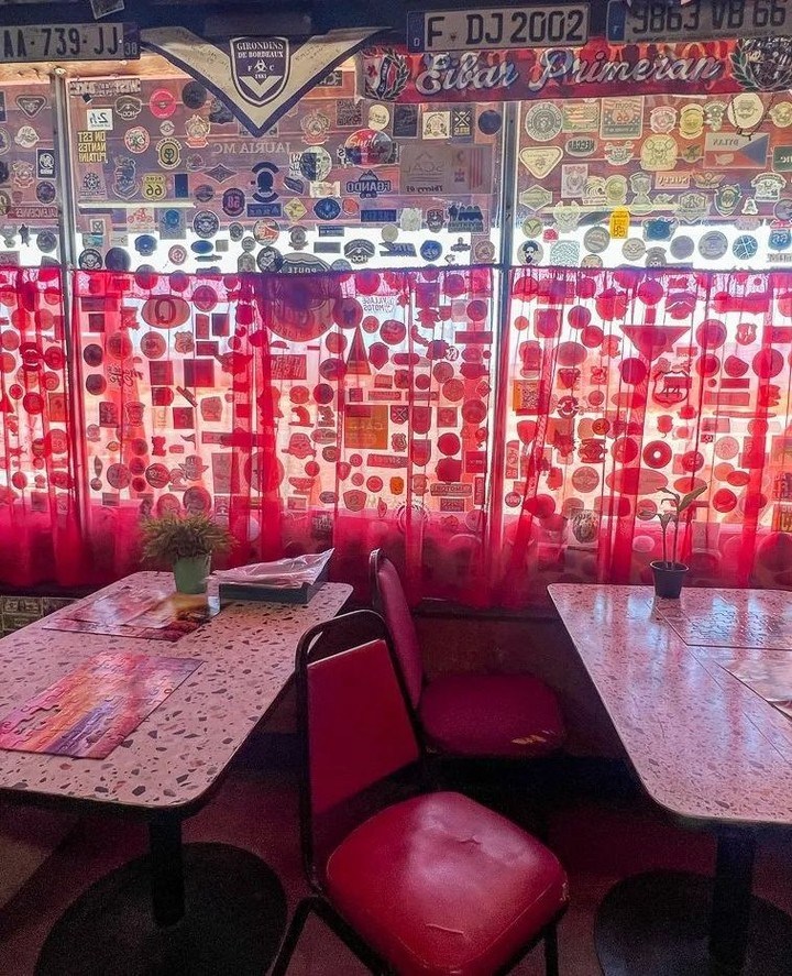 The interior of the Bagdad Cafe, with red curtains and windows covered in stickers, two tables with red chairs, and potted plants.