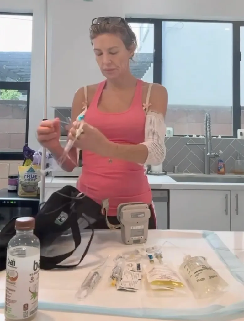 A woman in a pink top using an at-home IV drip, with medical supplies and an IV pump on a table in front of her.