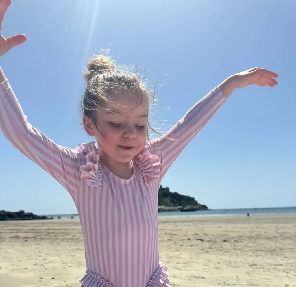 Sienna, a young girl with her hair in a bun, wearing a pink and white striped swimsuit with ruffles, stands on a sunny beach with her eyes closed and arms outstretched.