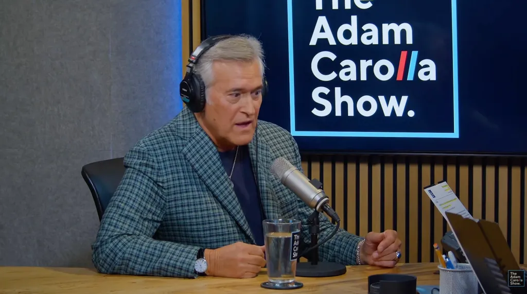 Bruce Campbell wearing headphones and a plaid jacket, speaking into a microphone with 