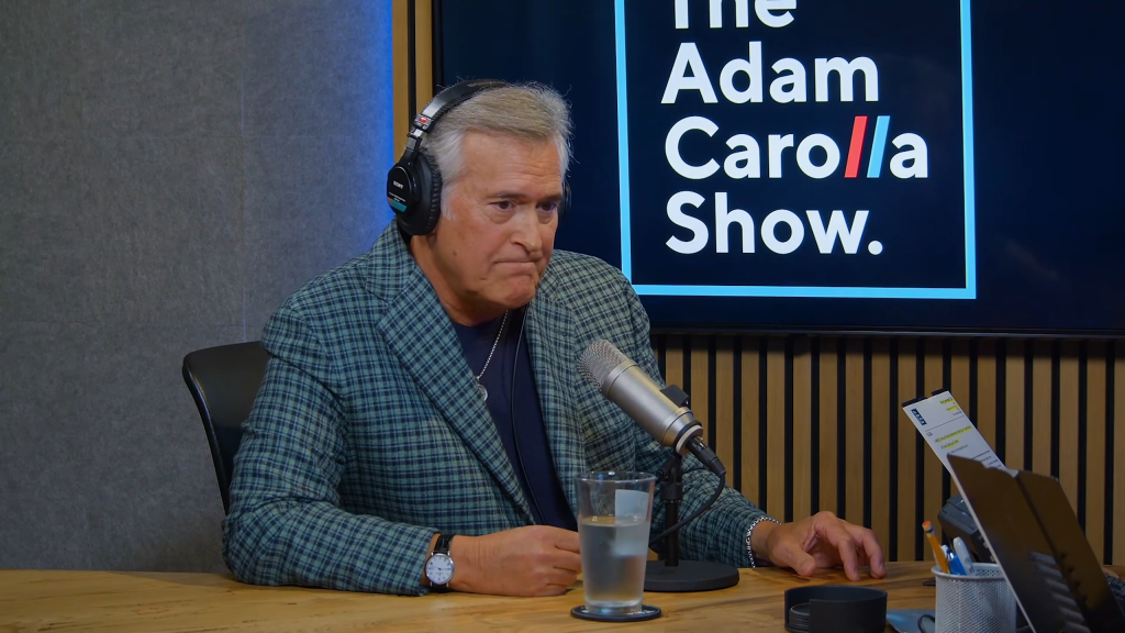Bruce Campbell wearing headphones and a plaid jacket sitting at a table with a microphone and a glass of water during 