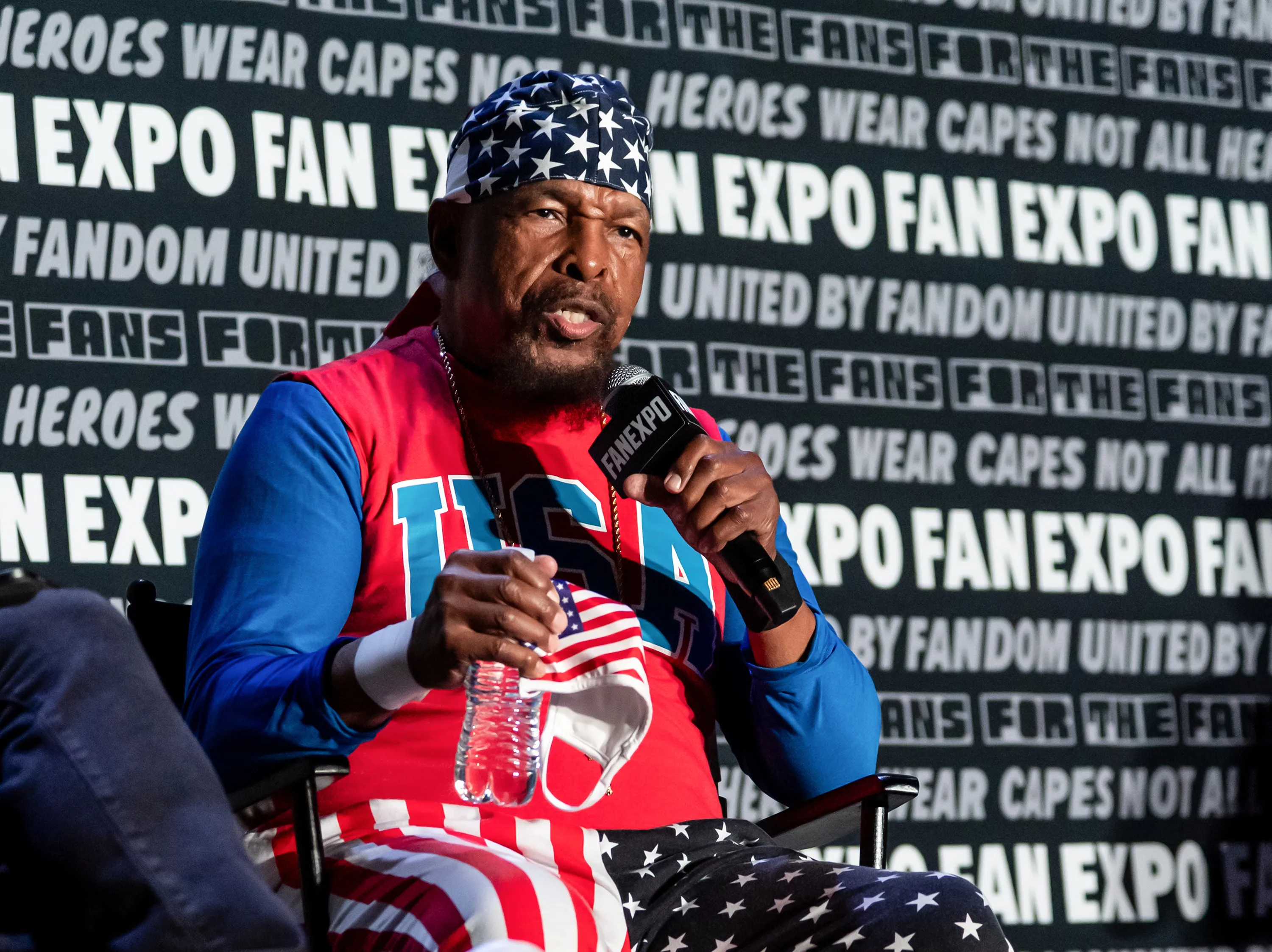 Mr. T, dressed in a patriotic American flag outfit, speaking into a microphone at a Fan Expo event.