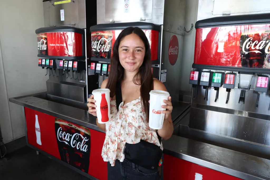 Seri holds two soft drinks in front of Coca-Cola fountain dispensers.