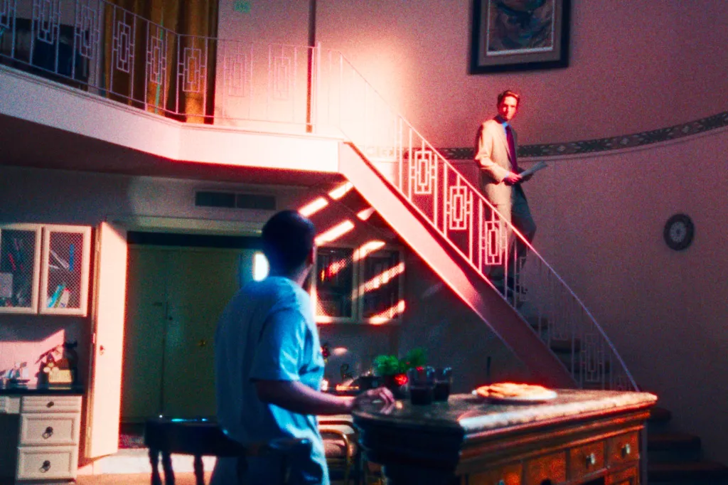 Tony Revolori stands facing away from the camera in the foreground, while Robert Pattinson descends a staircase in the background in a scene from 