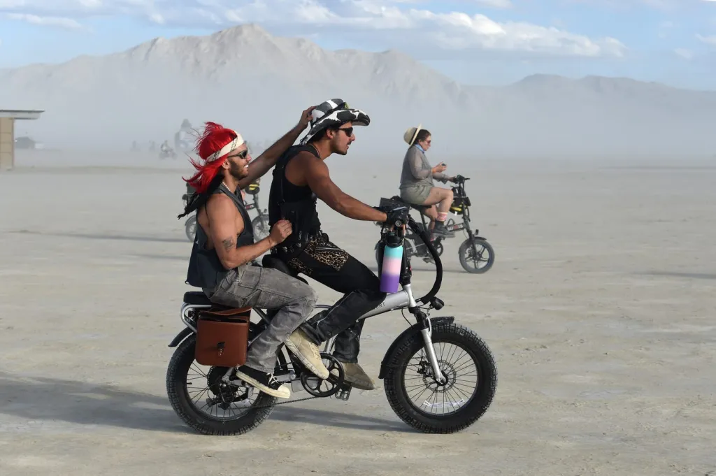 Two men on a bicycle at Burning Man, with another person on a bicycle behind them in the desert.