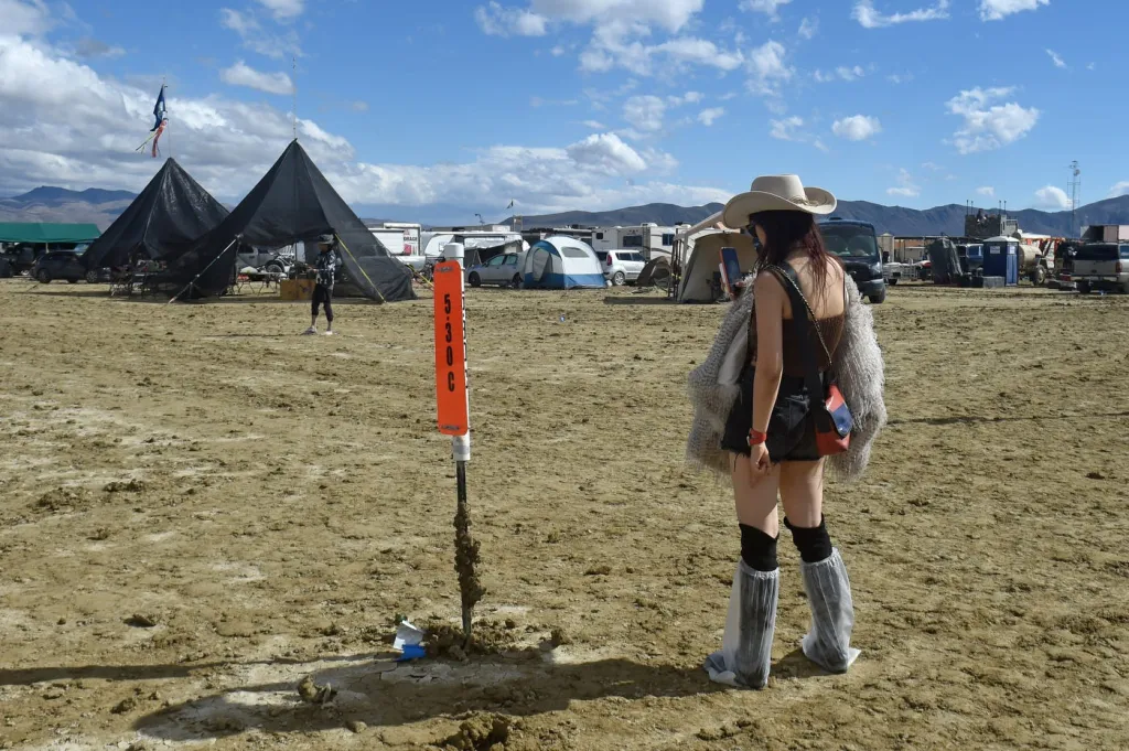 A woman in a cowboy hat and shorts looks at her phone in the dusty Black Rock Desert, with camping tents and vehicles in the background.