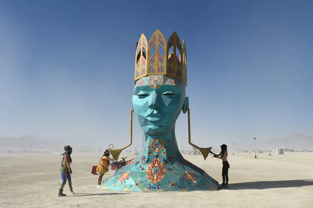 A large, blue, polygonal sculpture of a head with a golden crown and ornate patterns at its base stands in a desert with three people interacting with it.