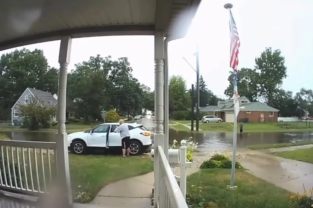 A person gets out of a white car parked on a lawn next to a flooded street.