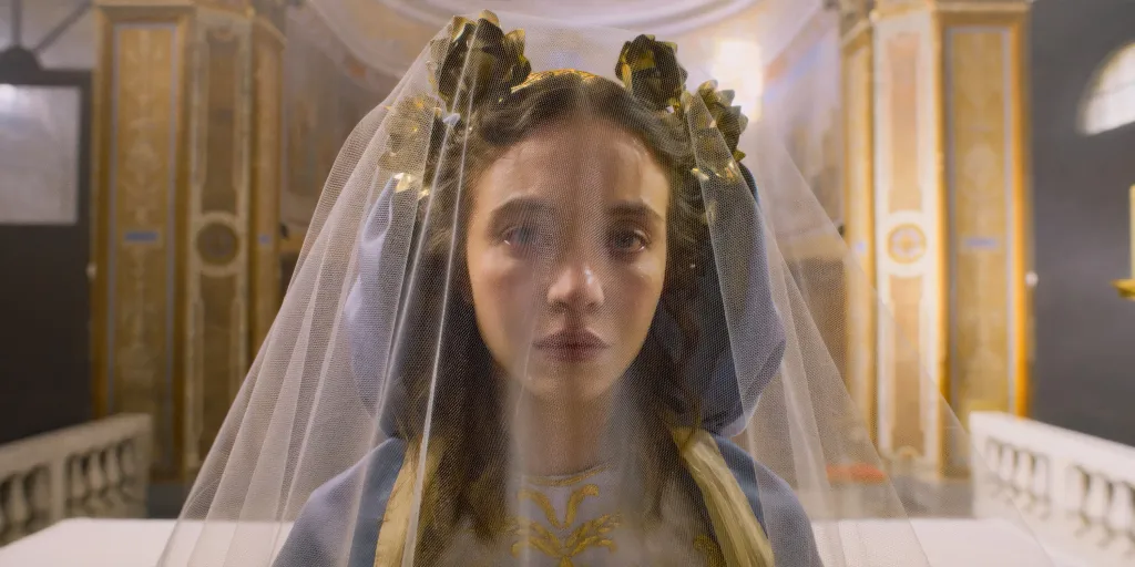 Sydney Sweeney, with dark hair and a gold leaf headpiece ,wears a light blue hooded dress with gold embroidery and a translucent white veil, looking forward with an unreadable expression.