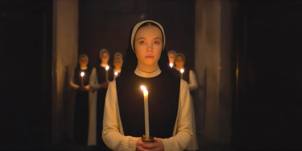 Sydney Sweeney as a nun, in a black and white habit and black dress with white shirt, holding a candle in a dark room with other nuns in the background from the movie Immaculate.