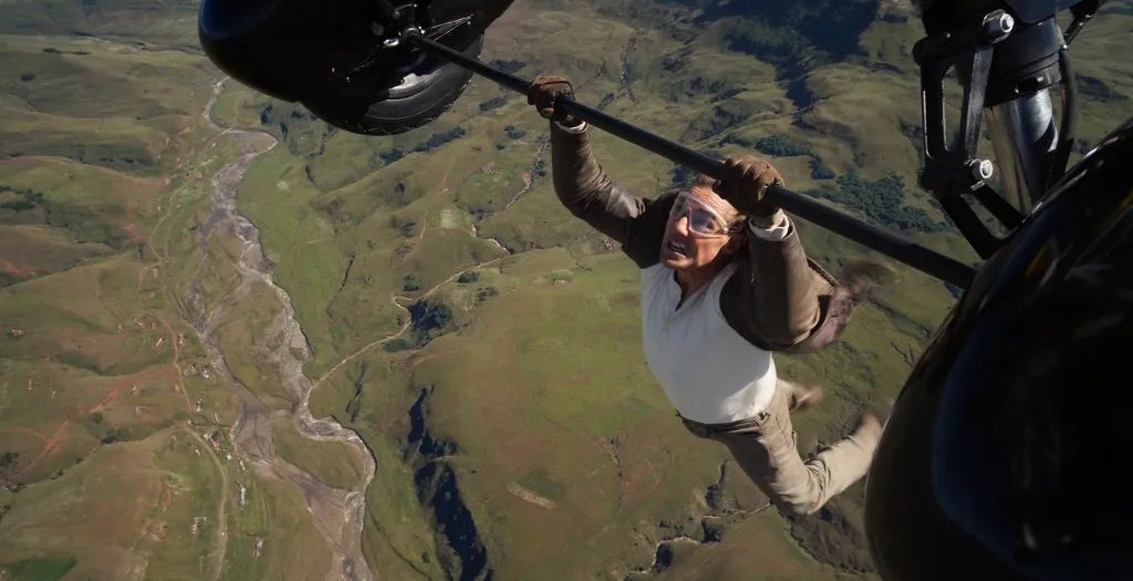 Tom Cruise, ni khaki pants, white T-shirt and a brown jacket, hanging from an airplane over a green valley.