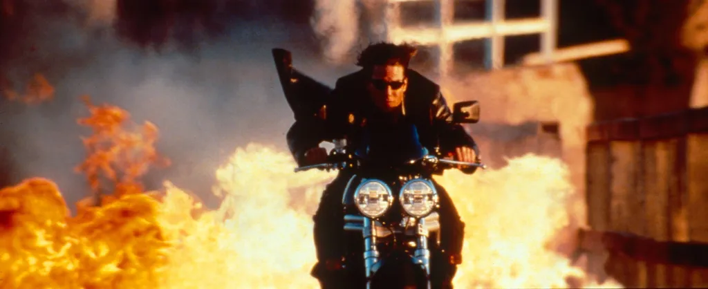 Tom Cruise, in a black leather jacket, on a motorcycle with flames exploding behind him in 
