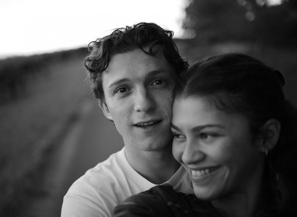 Black and white photo of Tom Holland smiling at the camera, with Zendaya smiling beside him.