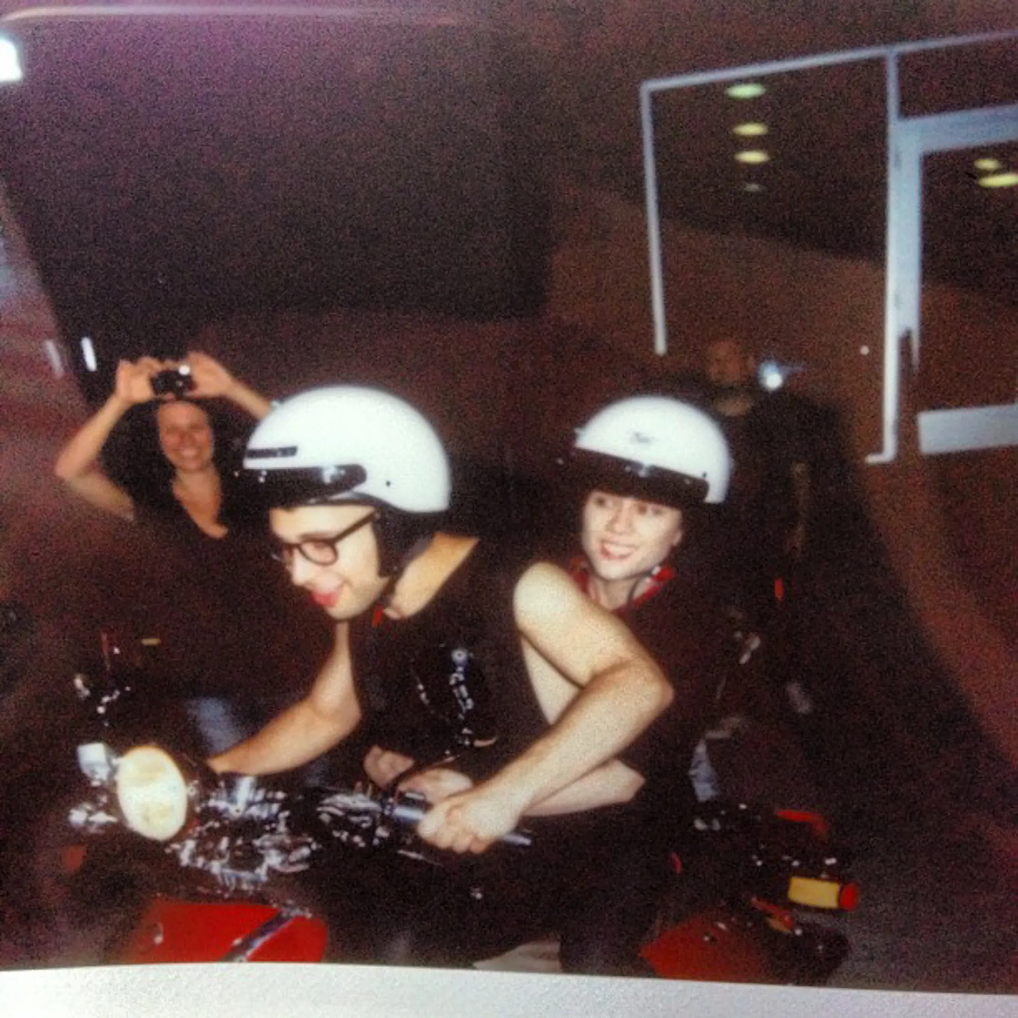 Jack Antonoff and Margaret Qualley on a motorcycle, both wearing white helmets, with a woman taking a photo in the background.
