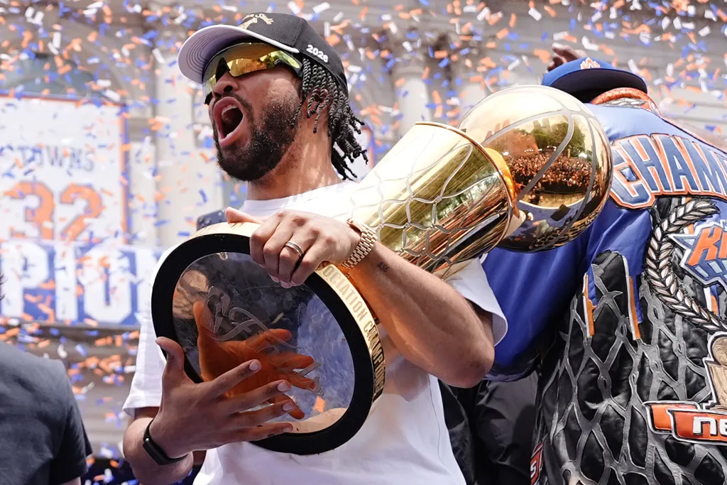 Jalen Brunson celebrates with the Larry O'Brien Championship Trophy amidst falling confetti.