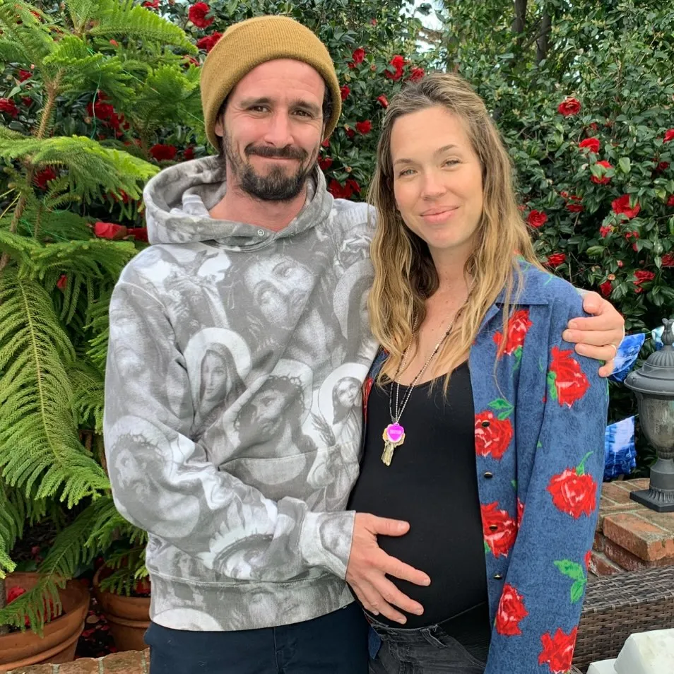 James Ransone and his wife Jamie, who is pregnant, stand together in front of a bush with red flowers.