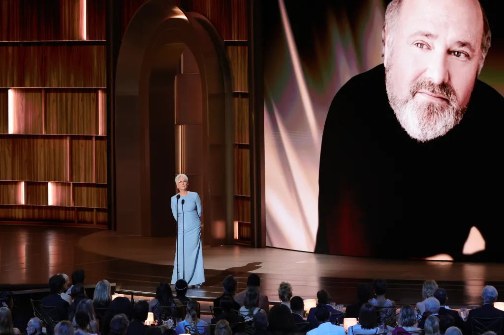 Jamie Lee Curtis speaks on stage at the 78th Primetime Emmy Awards with a large screen showing a man's face behind her.