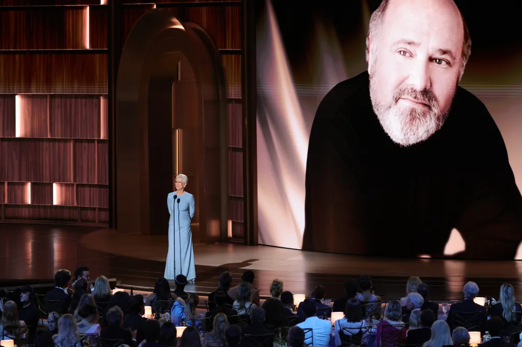 Jamie Lee Curtis speaks on stage at the 78th Primetime Emmy Awards, with a large screen behind her displaying an image of a man with a beard.