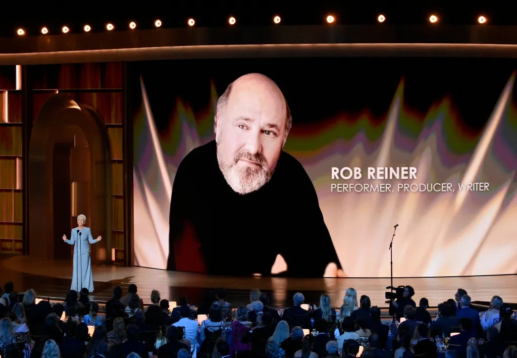 Jamie Lee Curtis speaks on stage at the Primetime Emmy Awards during a memoriam for Rob Reiner, whose photo and title appear on the screen behind her.