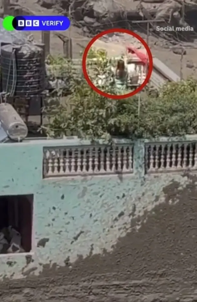 Three people on a balcony of a green house as floodwaters rage below.