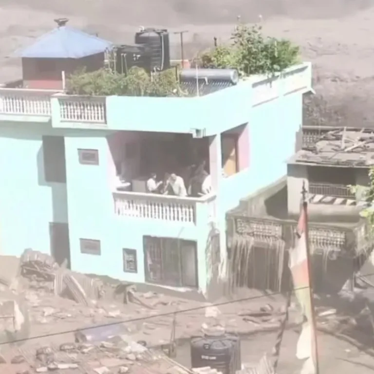 A green house stands intact amidst flood debris with three people on its balcony.