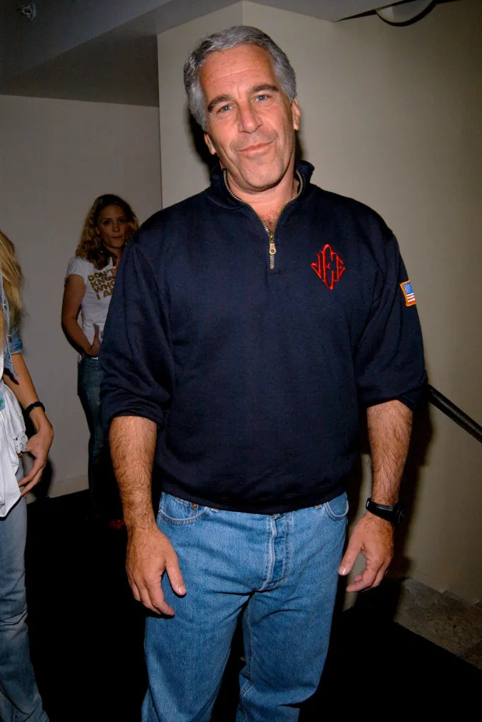 Jeffrey Epstein in a navy blue pullover with a red logo and an American flag patch, light blue jeans, and a black watch, standing in a hallway.