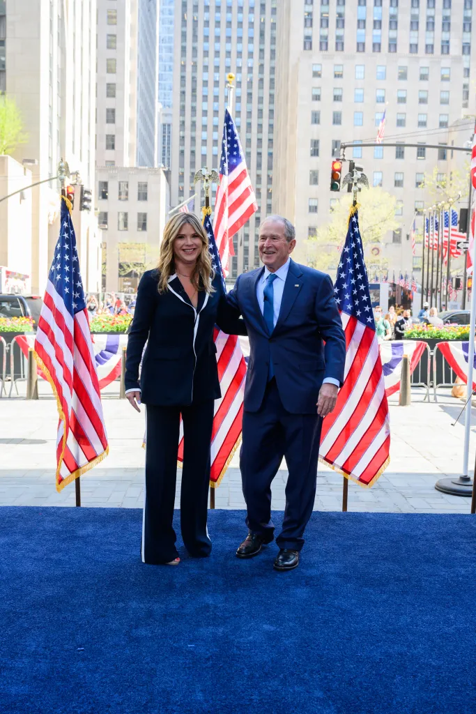 Jenna Bush Hager and George W. Bush smile, standing on a blue carpet with American flags around them, in an outdoor setting.