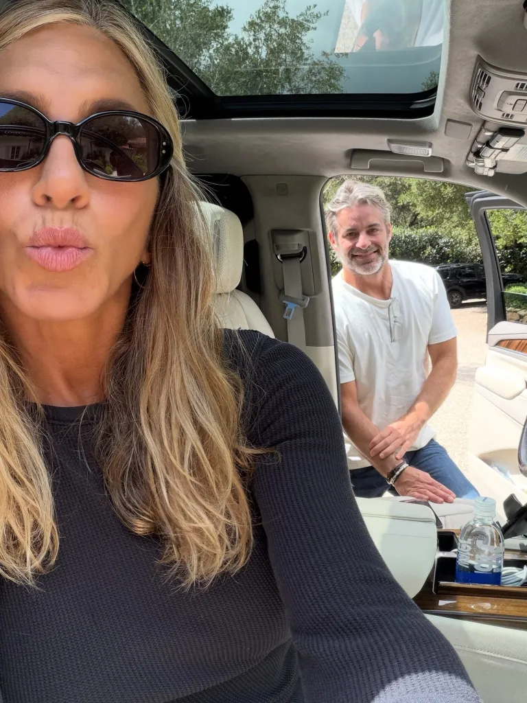 Jennifer Aniston takes a selfie in a car with a man smiling in the background.
