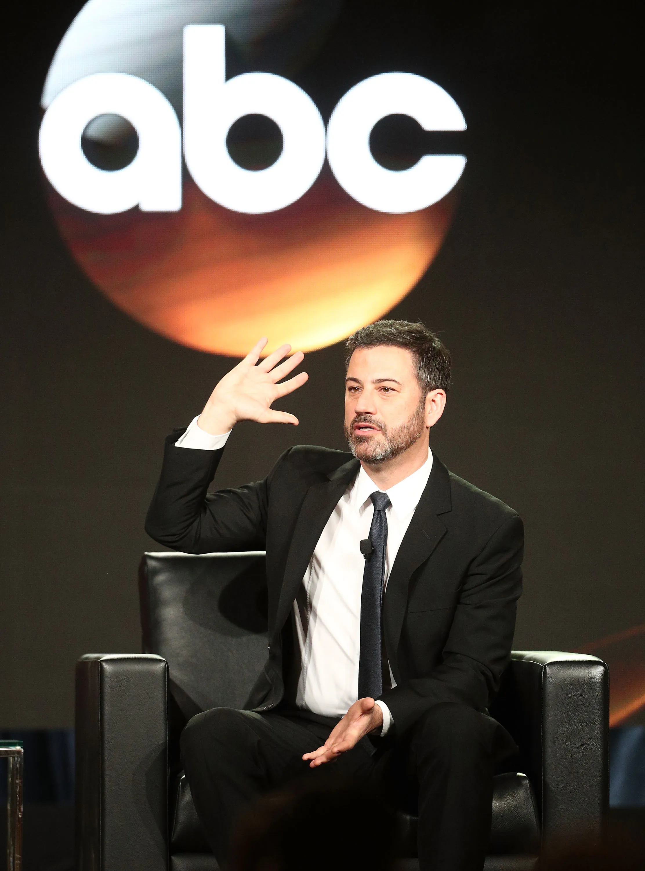 Jimmy Kimmel seated on a black chair, speaking with one hand raised, in front of a screen displaying the ABC logo.