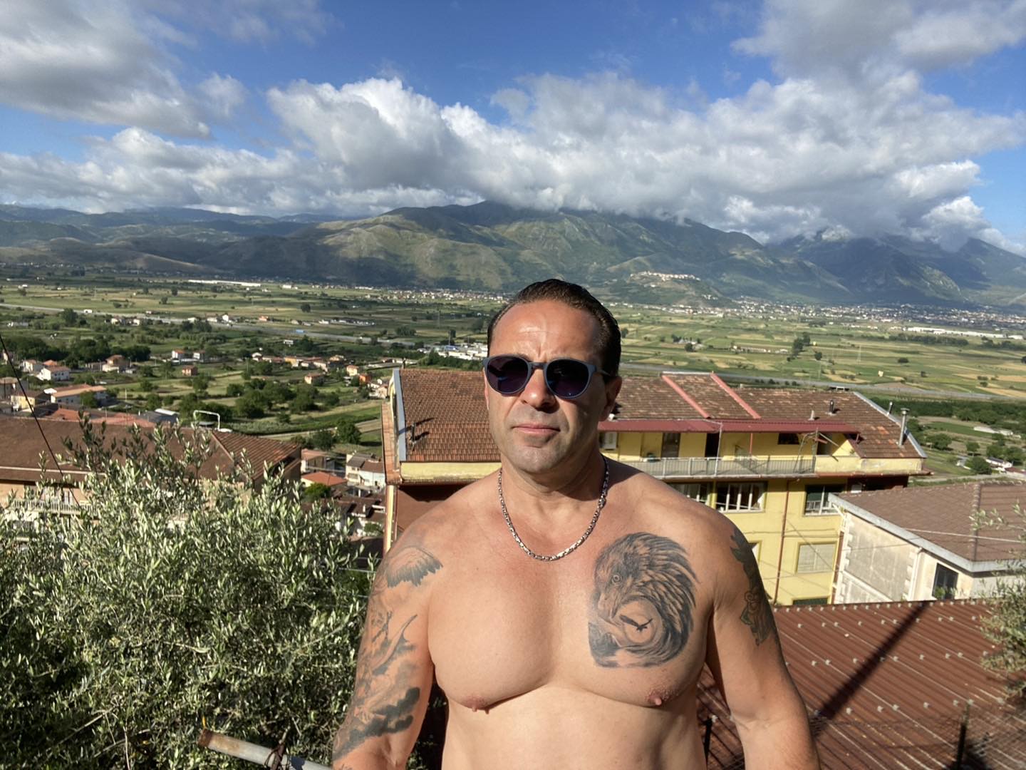 Joe Giudice shirtless wearing sunglasses and a silver chain, with a mountain range and valley in the background.