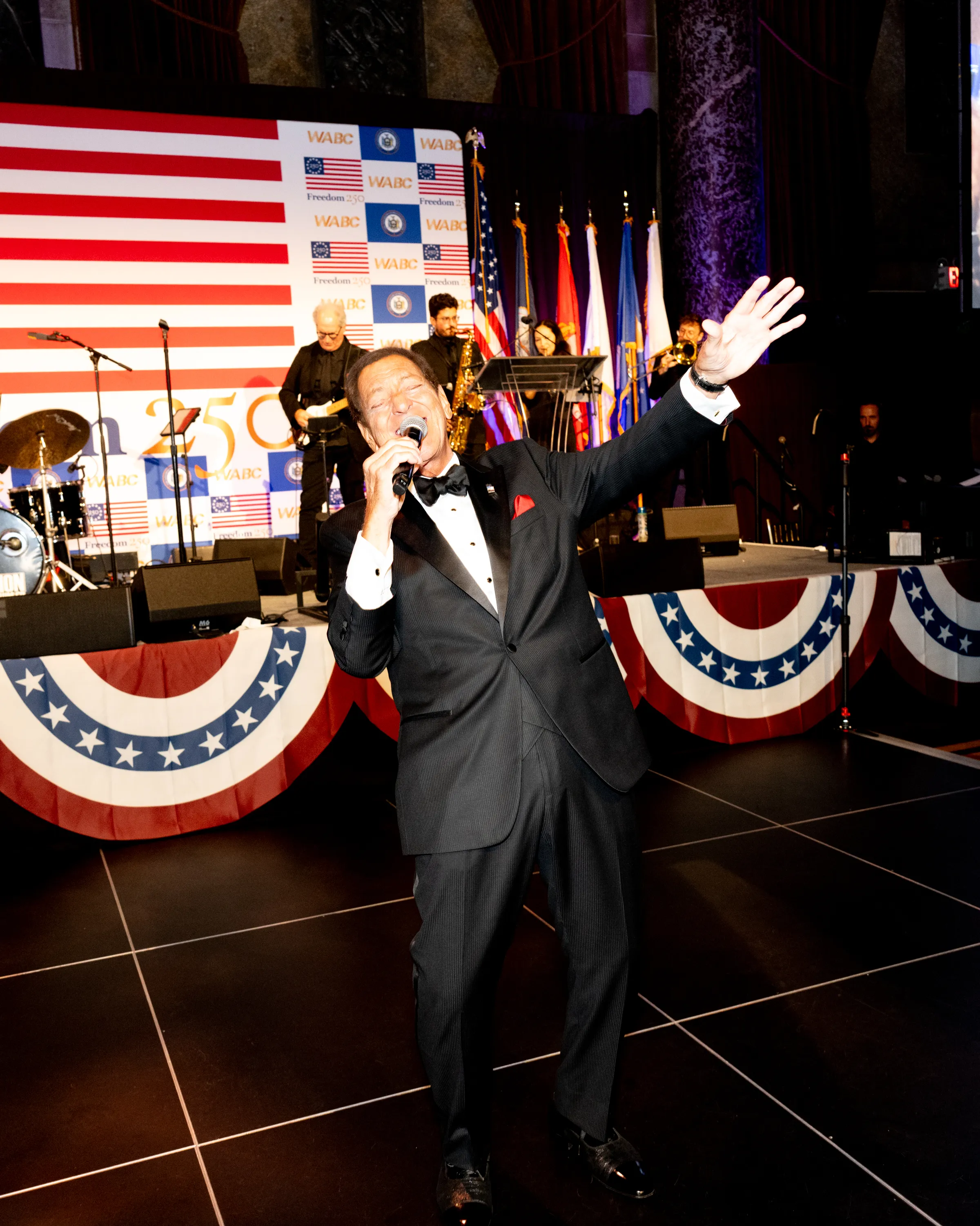 Joe Piscopo performing on stage, dressed in a tuxedo, at John Catsimatidis' 25th anniversary gala.