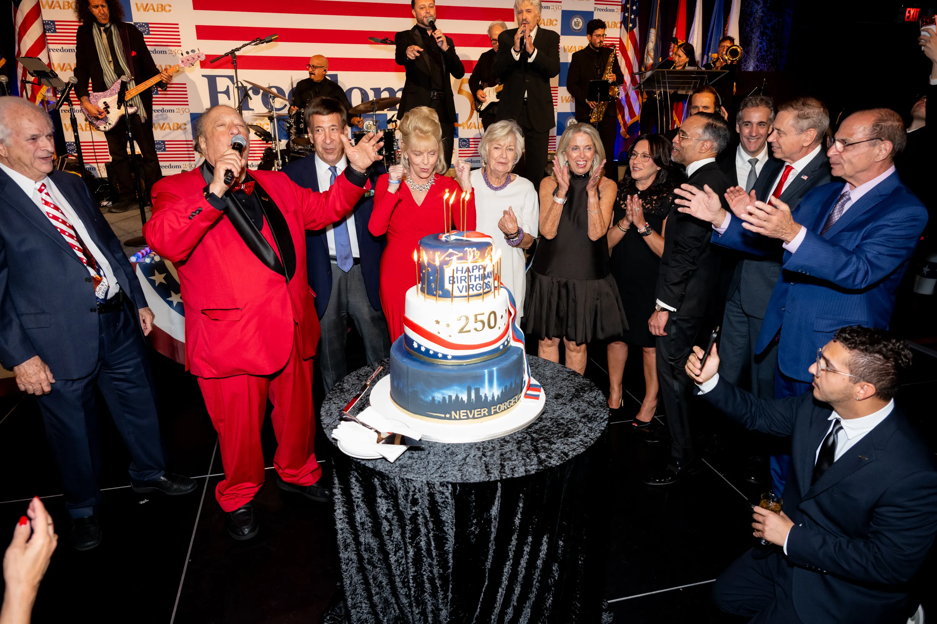 People gathered around a large birthday cake, some applauding, with a band in the background.
