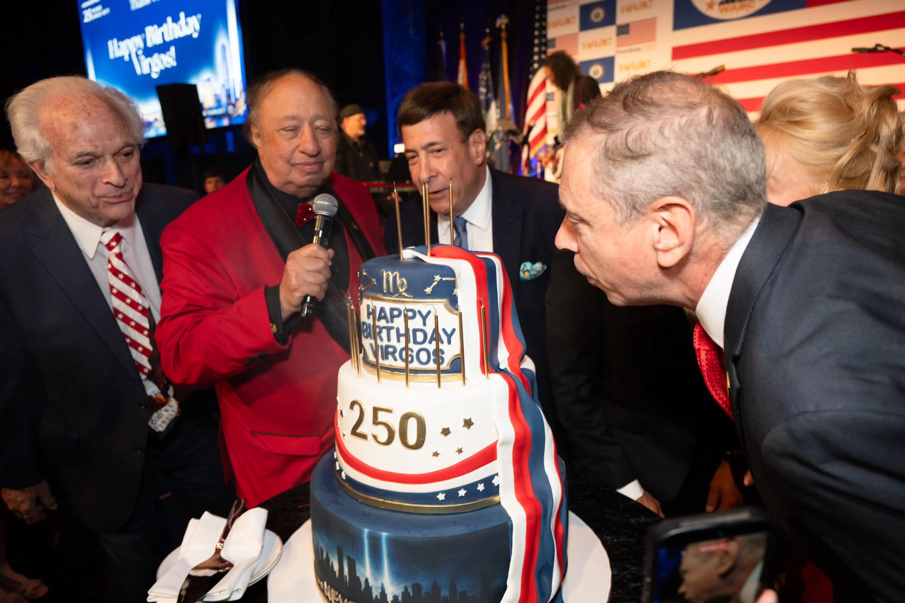 John Catsimatidis holding a microphone next to a birthday cake, as another man leans in to blow out candles.