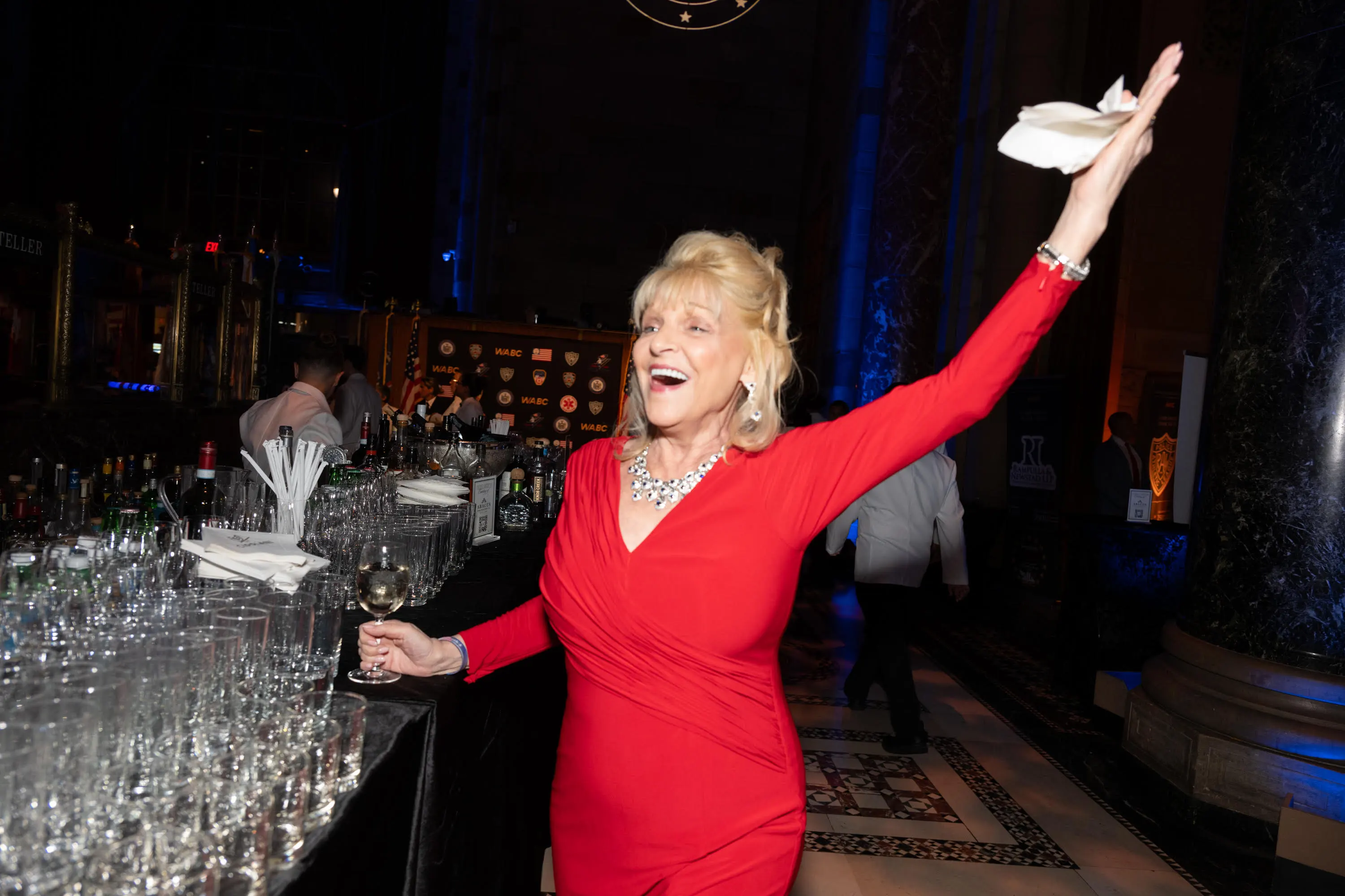 Margo Catsimatidis, dressed in a red gown and silver necklace, holds a glass of white wine and raises her other hand in celebration at a gala.