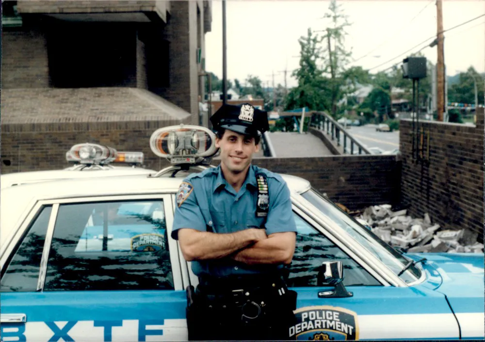 NYPD ESU Officer John D'Allara in uniform, standing with arms crossed in front of a police car.
