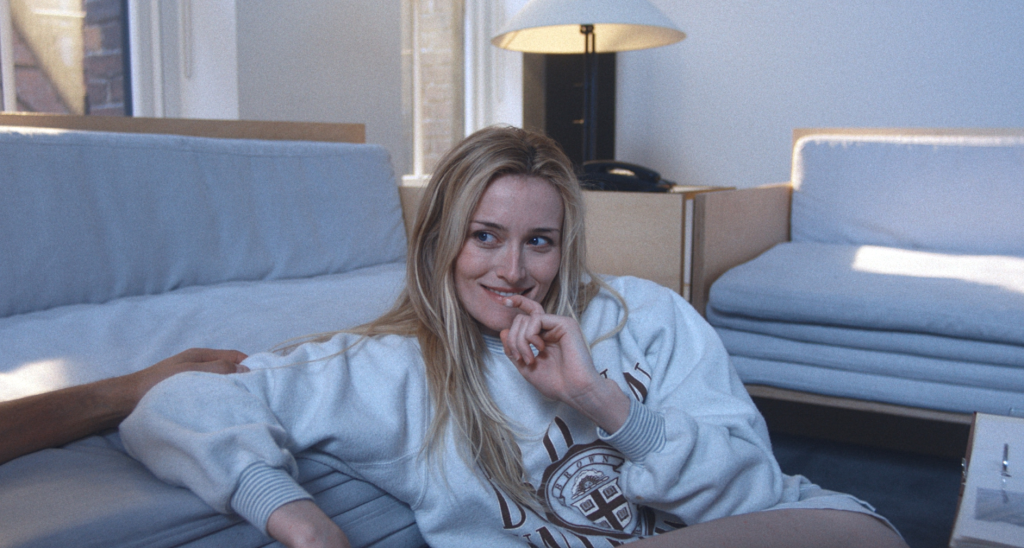 Sarah Pidgeon as Carolyn Bessette, smiling while resting on a sofa.