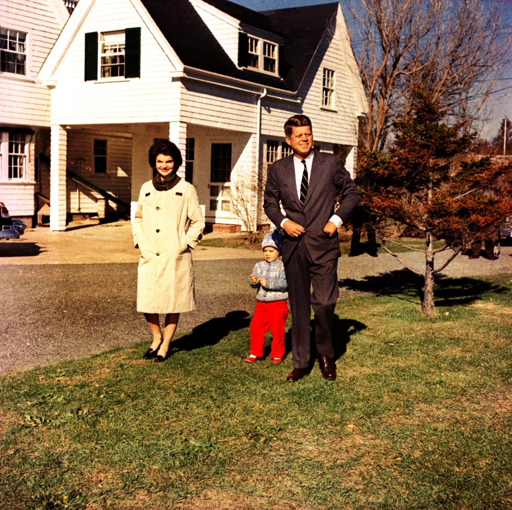 John F. Kennedy, Jacqueline Kennedy, and their daughter Caroline, walking outdoors in front of a white clapboard house.