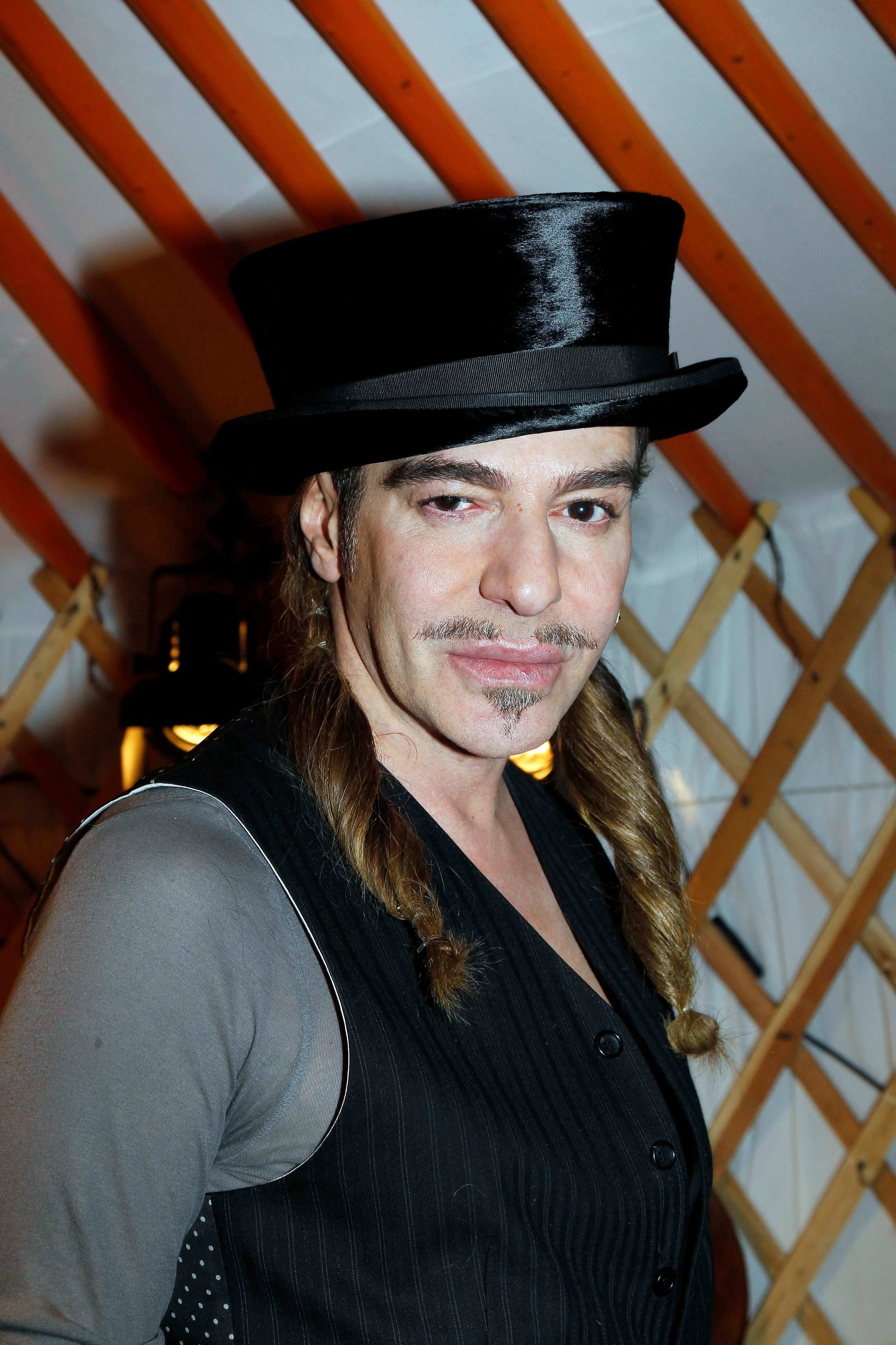 John Galliano wearing a black top hat, pinstriped vest, and gray shirt.