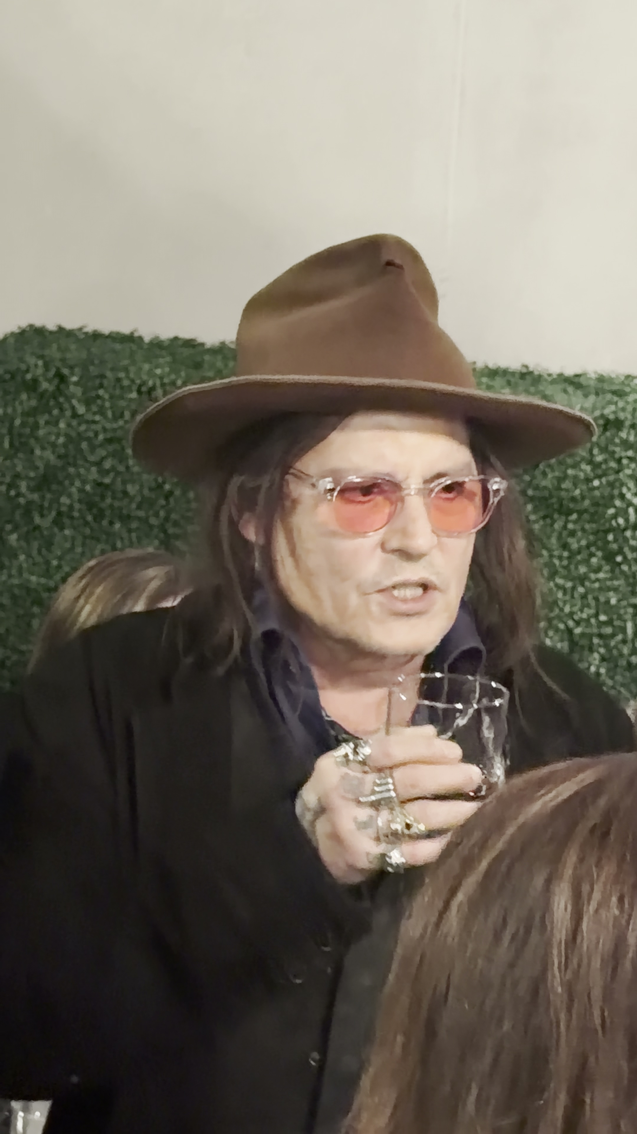 Johnny Depp in a brown hat and pink glasses, holding a glass and speaking in front of a green artificial plant backdrop.