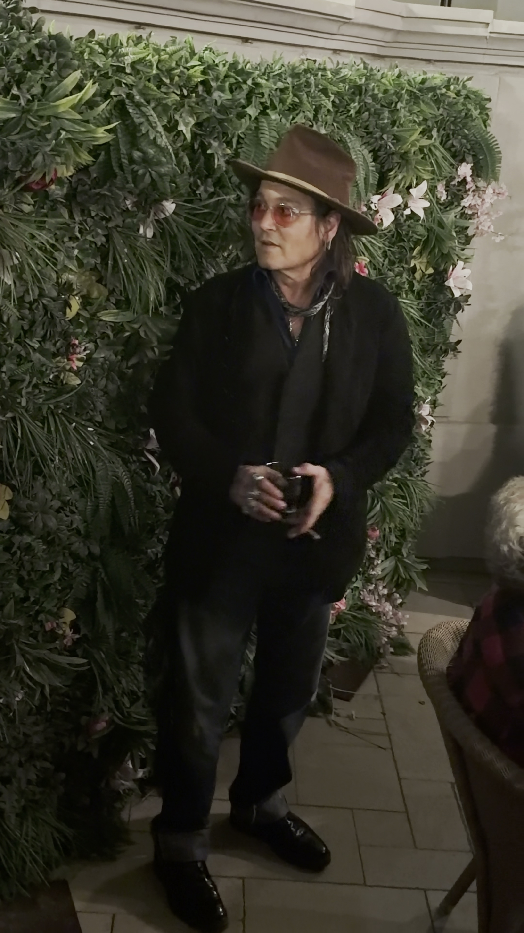 Johnny Depp wearing a brown fedora, sunglasses, and a black outfit stands in front of an artificial plant wall, holding a drink.