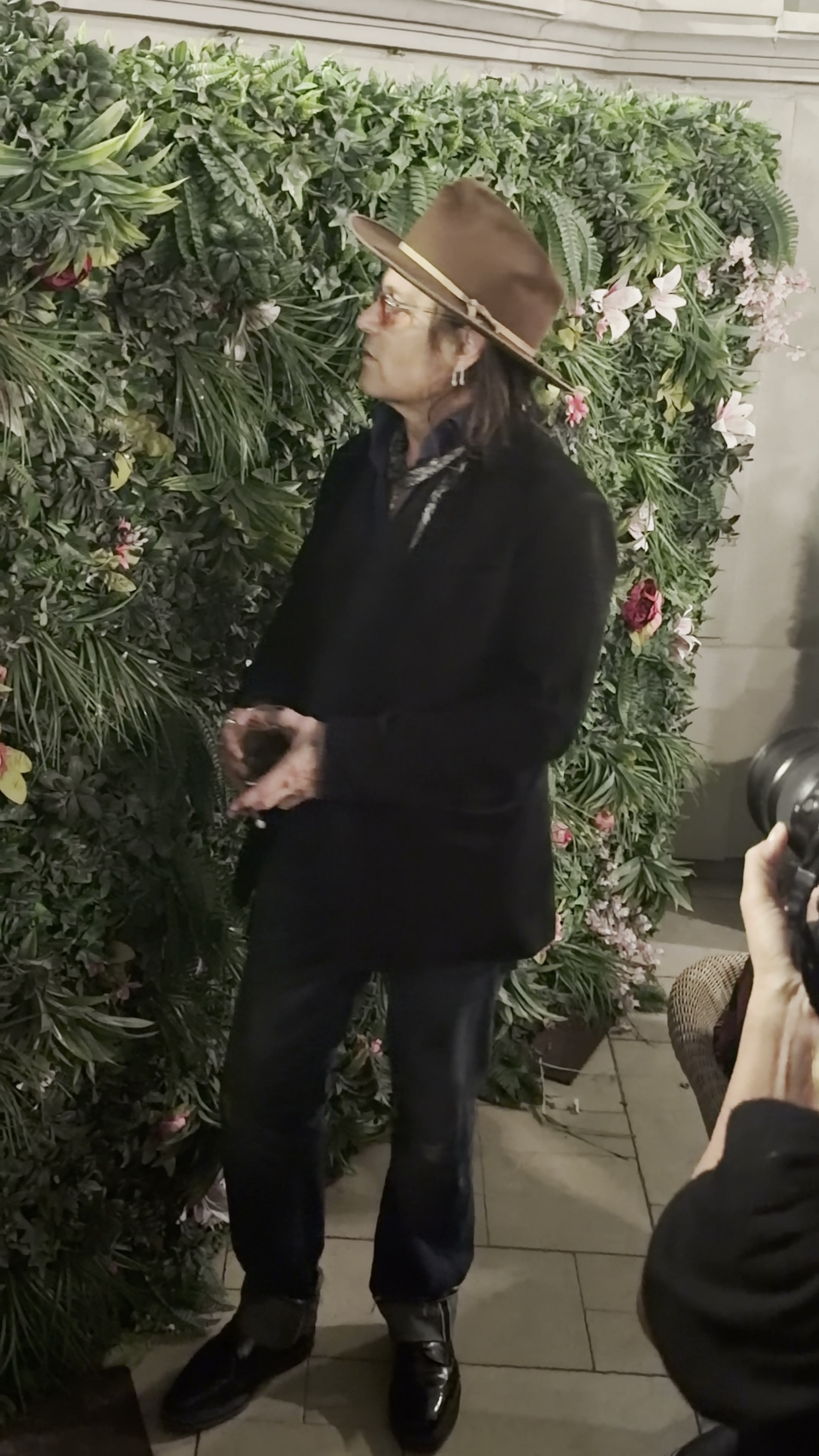 Johnny Depp in a black jacket and brown hat stands in front of a green artificial plant wall, with a photographer visible at the bottom right.