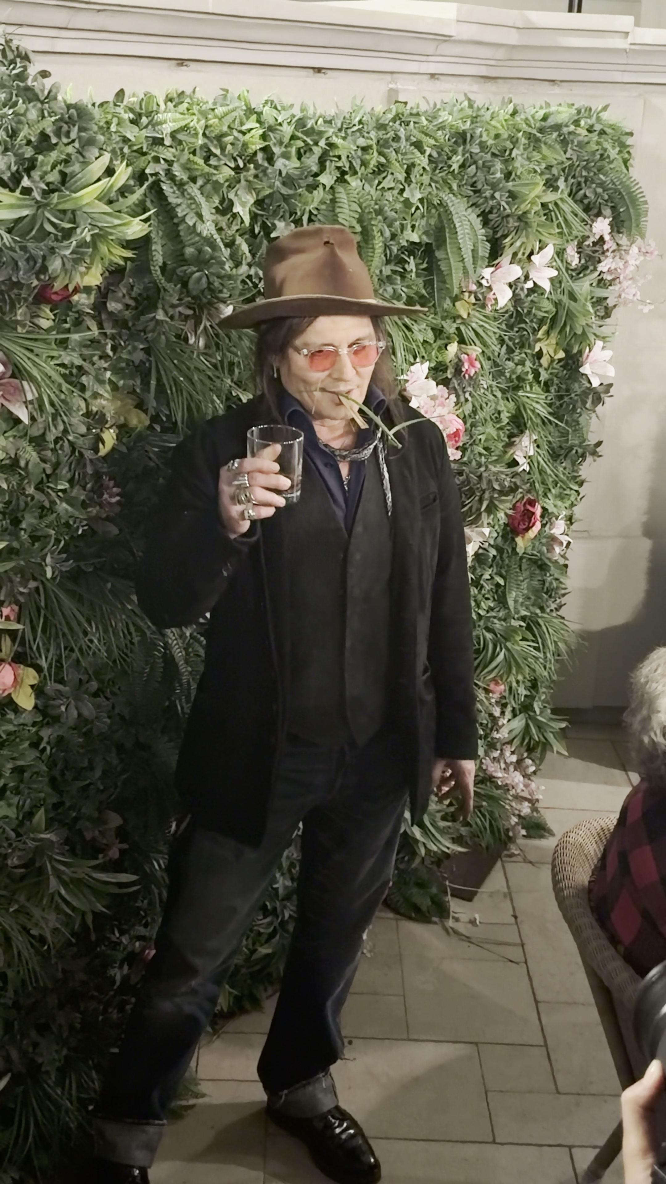 Johnny Depp in a brown hat and red-tinted glasses holding a drink and chewing on an artificial plant.