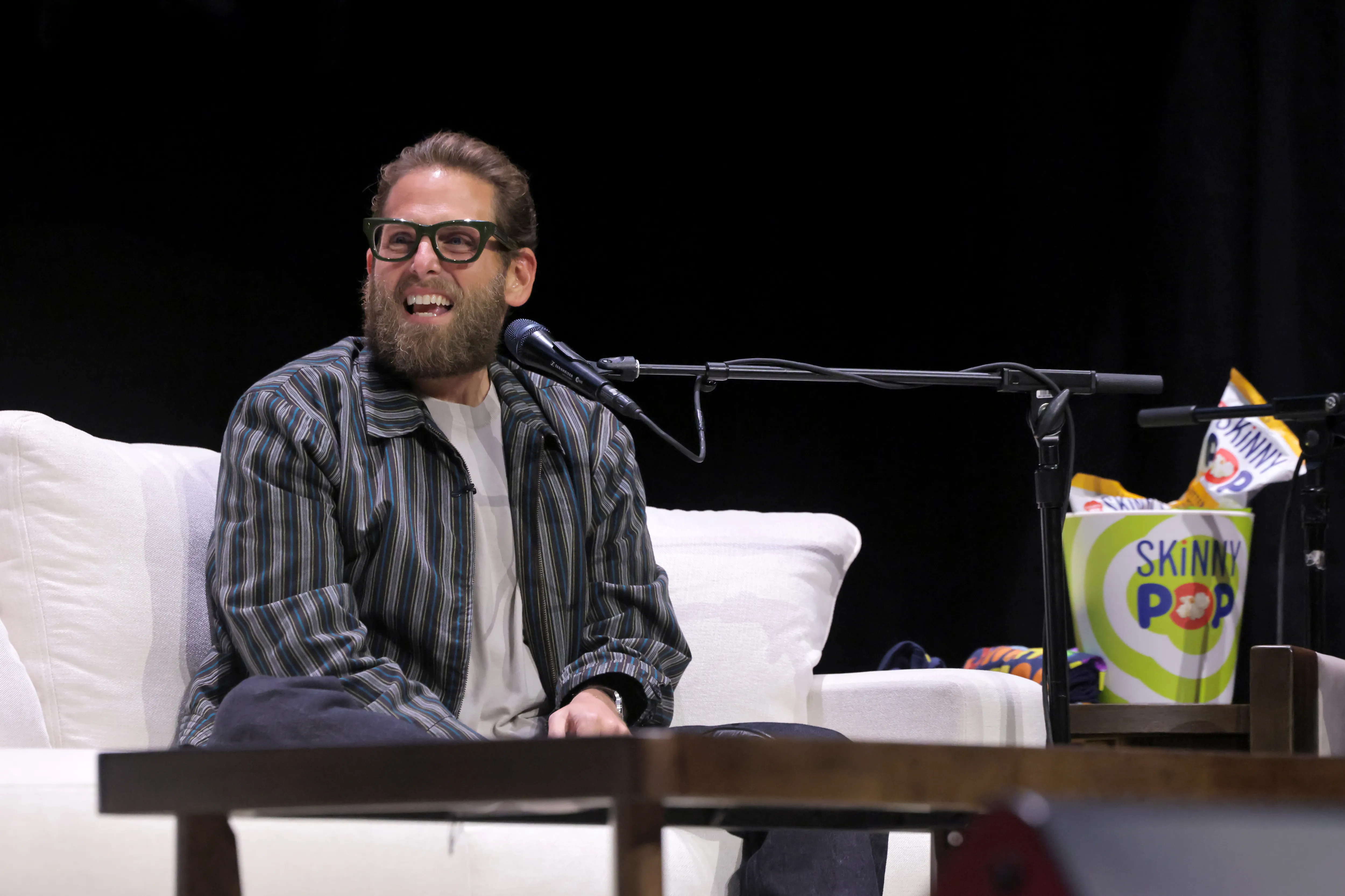 Jonah Hill, wearing glasses and a striped jacket, smiles broadly while sitting on a white sofa with microphones in front of him.