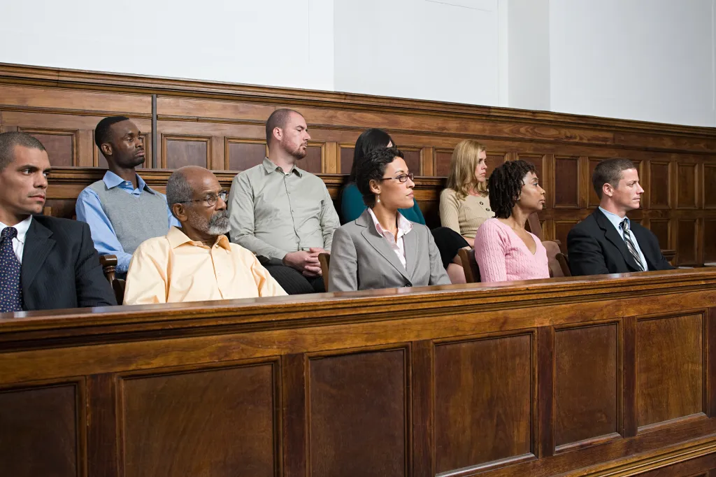 A diverse group of jurors, men and women of varying ages and ethnicities, sit in a jury box.