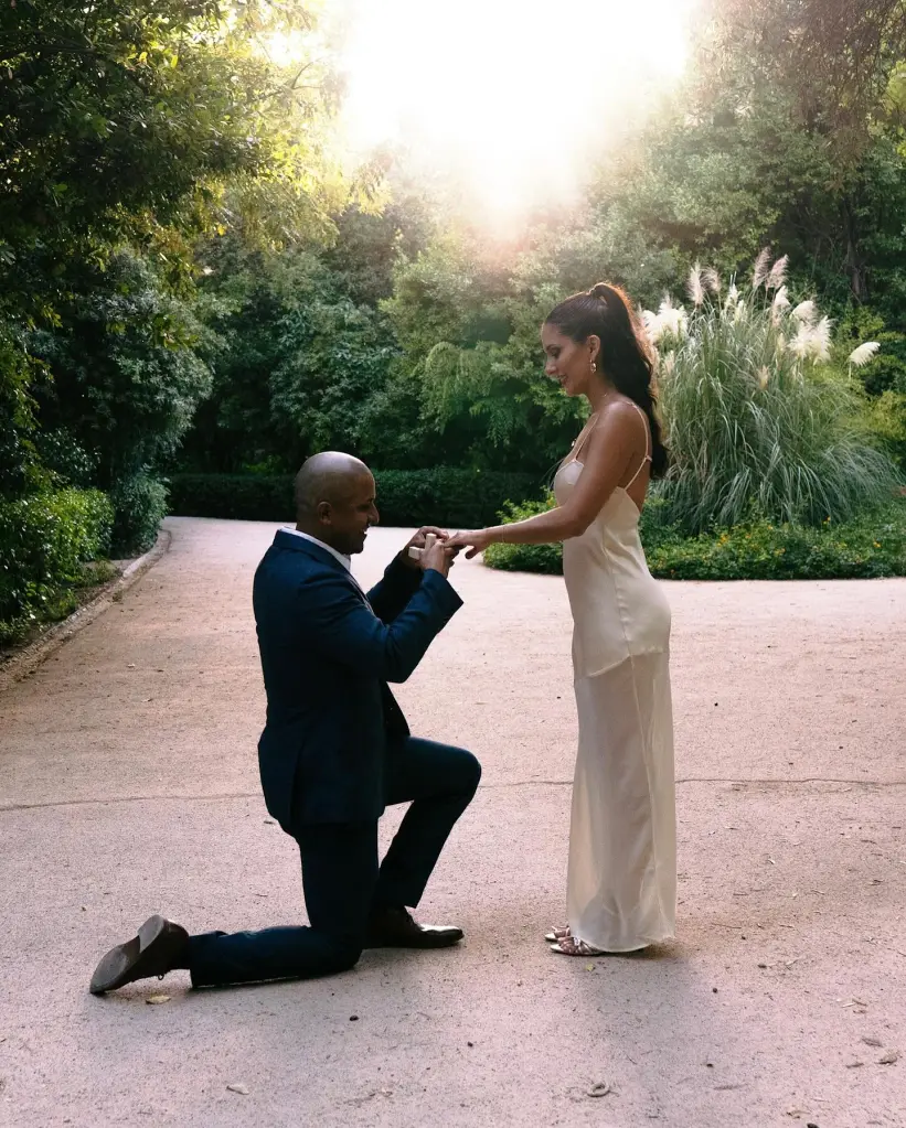 A man proposes to a woman on a pathway surrounded by trees and plants.