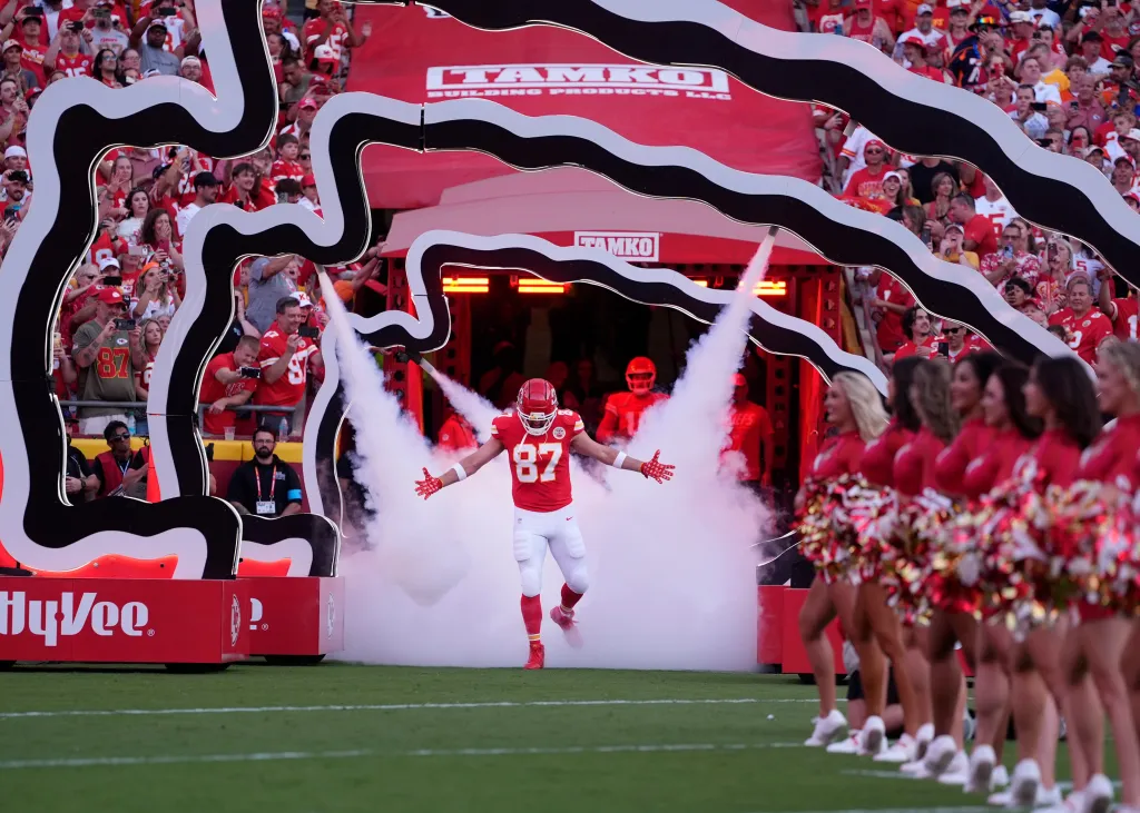 Kansas City Chiefs tight end Travis Kelce (87) emerges through smoke from a tunnel onto the field, flanked by cheerleaders.
