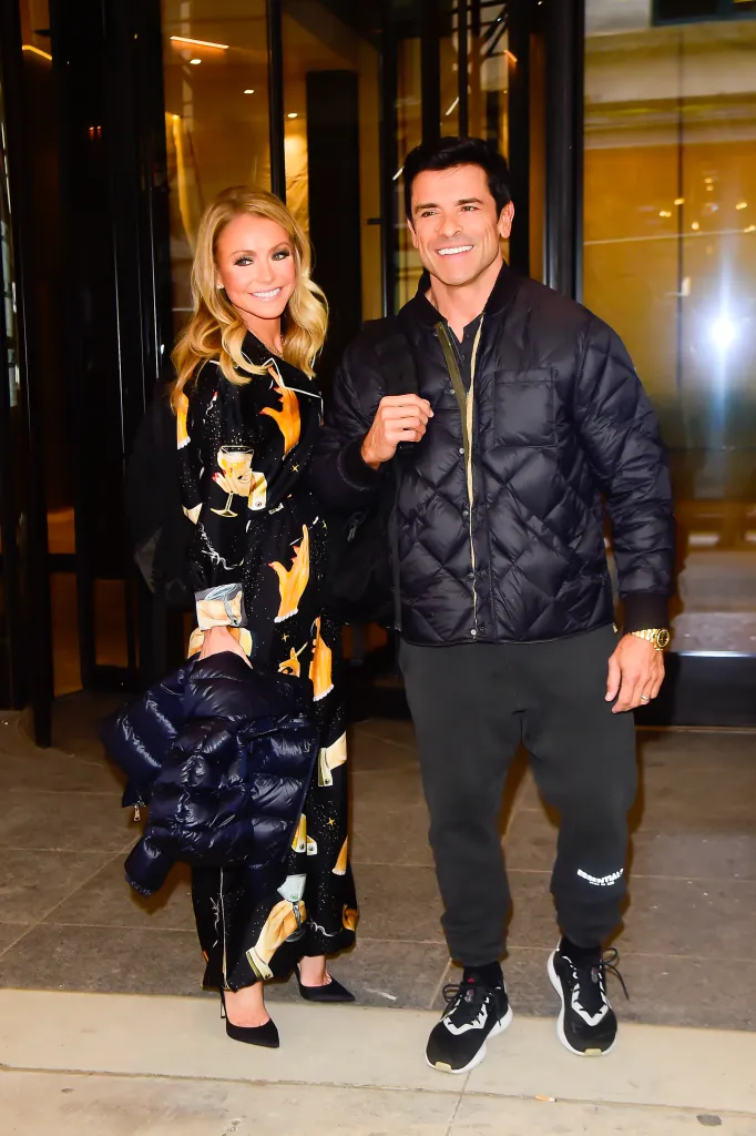 Kelly Ripa in a black patterned shirt dress and Mark Consuelos in a black puffer jacket and sweatpants smiling.