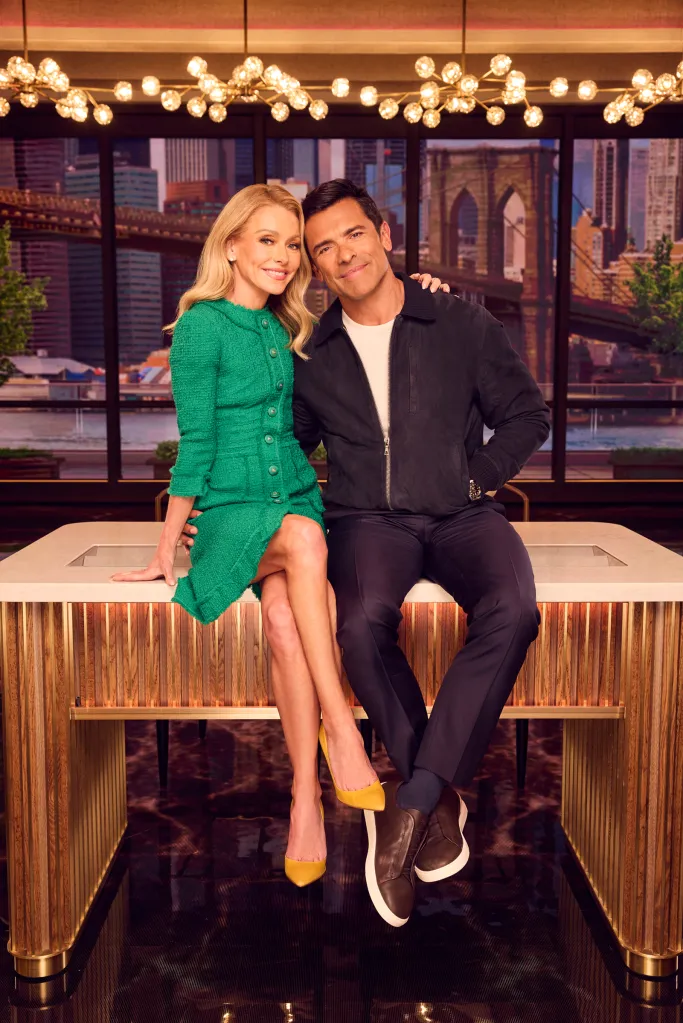Kelly Ripa and Mark Consuelos pose while sitting on a desk.