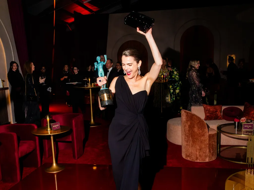 Keri Russell, in a black gown, holding an Actor Awards trophy above her head.
